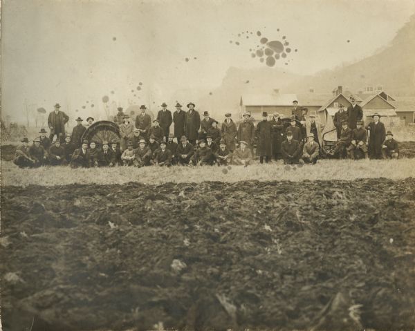 View across plowed field of group of employees of La Crosse Tractor? The men are wearing long jackets, neckties, gloves, and hats. In the background are farm or industrial buildings, and a bluff on the right.