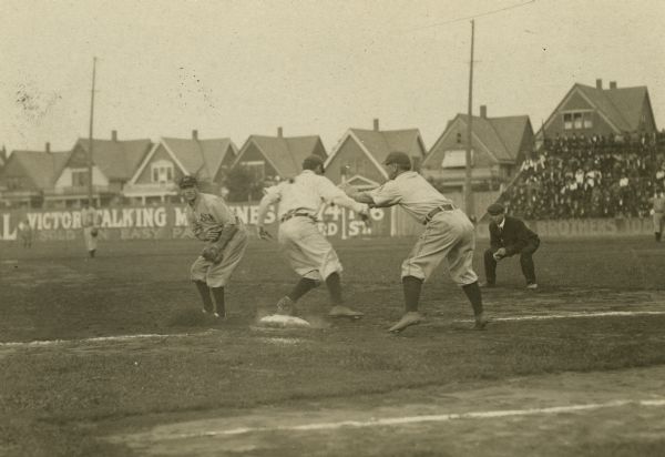 Runner reaching first base with coach signaling for him to stop. Defensive players and the umpire look on. A portion of the crowd in the stands is visible on the right. A row of houses can be seen over the outfield fence, which has a sign that reads, in part: "Victor Talking Machines."