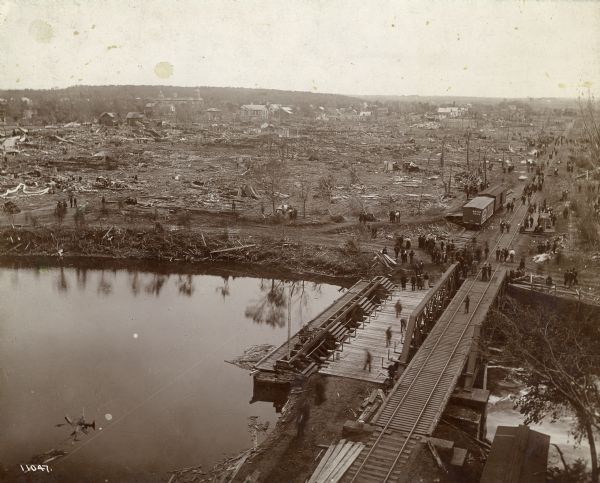Elevated view of New Richmond after the tornado hit on June 12, 1899. Many people are gathered around the railroad tracks and a few others are seen among the ruins of homes and businesses. The Willow River is visible in the foreground.
