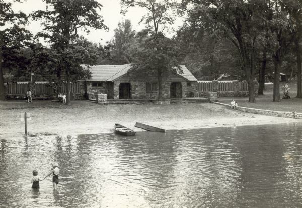 View of the new bath house at Devil's Lake State Park, taken from the pier. Two small boats are pulled up to the shore and two girls are wading in the water on the left.