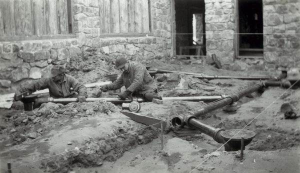 An interior view of partially-built dressing court, showing men working at roughing in plumbing to floor drains of the bathhouse at Devil's Lake State Park.