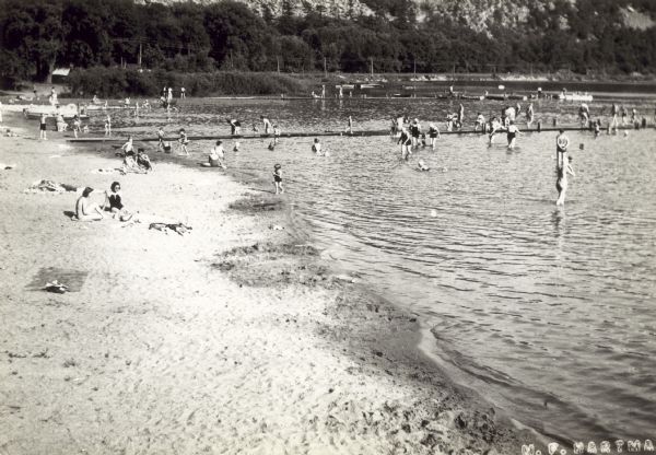 Men, women, and children swimming, playing, and relaxing at the beach at Devil's Lake State Park. Two long docks extend into the water and the scene is reportedly in front of the bath house.