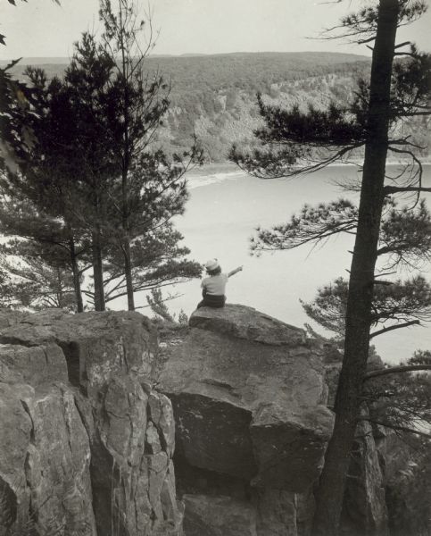 A vista of Devil's Lake from the high rock formations at Devil's Lake State Park. A girl is posed in the center on a rock with her back toward the camera, pointing out toward the lake.