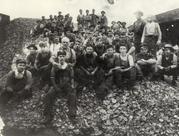 A large group of men of various ages sitting outdoors on a large mound of empty clam shells. The shells have holes drilled in them, probably for the pearl button factory. Most of the men are wearing overalls, and some wear hats.