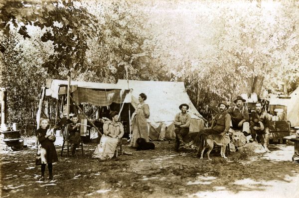 Clam camp housing and tents with forest in the background. Four men are sitting on the right, in the center are two women, one standing and one sitting, and on the left are two children. There are two dogs and a stove is on the far left side.