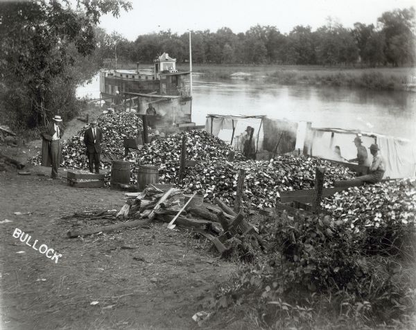 Elevated view of six men on the shore of a river, with piles of clam shells inside loose wooden fences near a pile of firewood and an axe. The firewood may be to provide heat for steaming the clams. A boat is on the river in the background. The two men standing on the left are wearing suits, ties, and hats, while the other men are in work clothes.