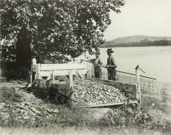Two men stand on the banks of a river near a mussel fishery, probably along the Mississippi River. The far shoreline of the river is in the background. The shells were mainly used for the manufacture of pearl buttons.