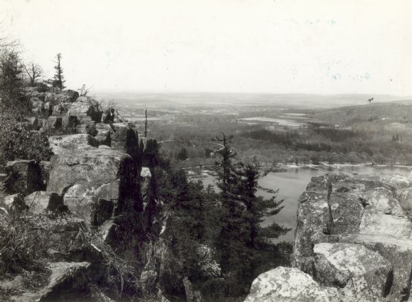 A scenic view of the south end of Devil's Lake State Park with the bluffs in the foreground, a view of the lake, and the surrounding area in the background. On the left side of the image is a man in a suit coat and hat sitting on a bluff.