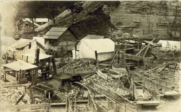 Elevated view of clam camp with tents, clam shell debris, and wooden buildings. A bluff rises up behind the buildings.