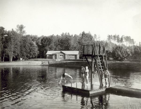 Two boys and two girls swimming at the Bad River at Copper Falls State Park. One of the girls is diving off the dock that also has a platform. In the background on the other side of the river is a building, probably the bathhouse.