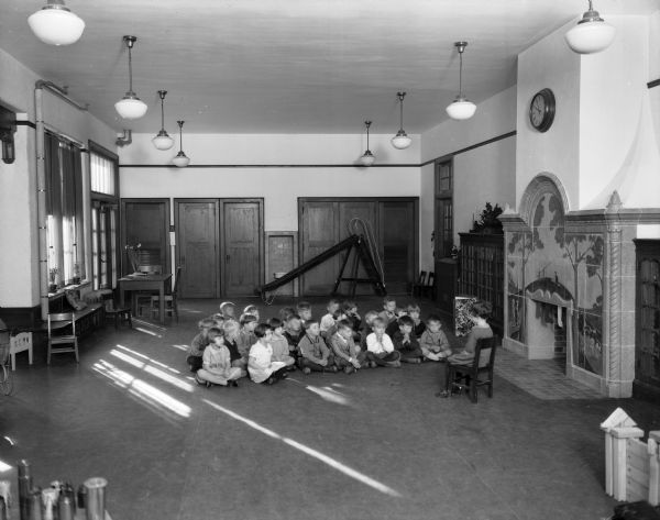 A group of children sit cross-legged on the floor of the kindergarten room at Lowell School. Dorothy Browne is the child sitting in the chair facing them. There is a ceramic tiled fireplace near them on the right, surrounded by bookcases. In the background is a slide.