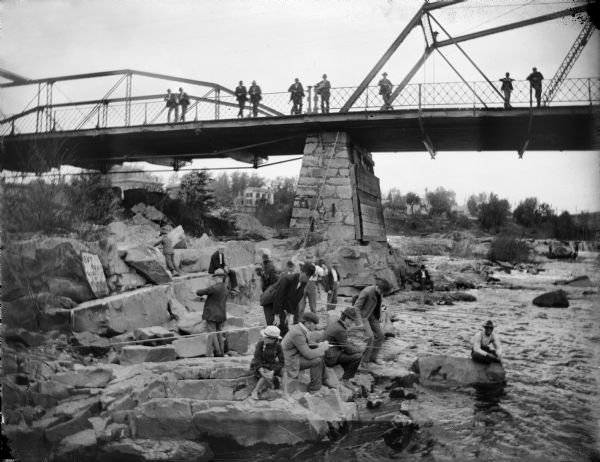 View along shoreline of group of men and boys fishing from rocks under a bridge, while others watch from above. Houses are in the far background on a hill. A sign for a grocery is propped on a rock on the left.