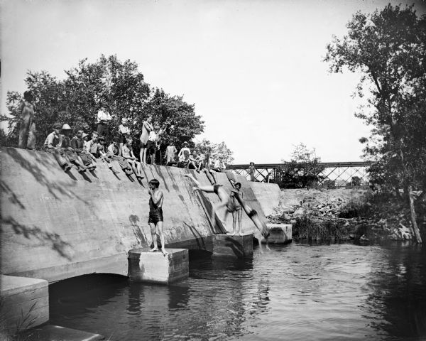 A large group of boys are standing and sitting on the bulkhead of a powerhouse before a flood. The boys could dive under one side of the structure and swim to the other side, as two of them are doing.