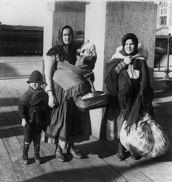 Two immigrant women in headscarves pose with a baby and a young boy. They are both holding baskets and bundles, and are standing on a platform.