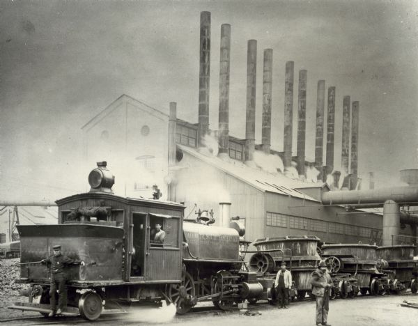 Men pose on and around a large steam engine locomotive and mineral cars(?) in front of a factory with tall smokestacks. The side of the engine reads: "Calumet & South Eastern R.R."