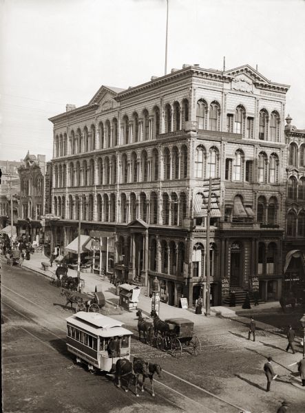Elevated view of building on corner in downtown area. There are horse-drawn vehicles in the street, and pedestrians on the sidewalk and crossing the street.