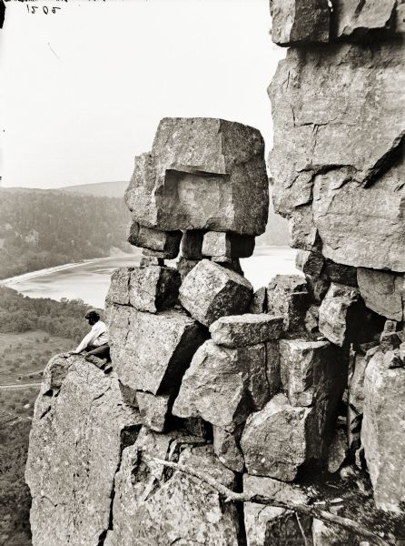 Devil's Lake Vicinity; boulder on top of a cliff at the foot of Devil's Doorway. There is a man sitting on a rock ledge.