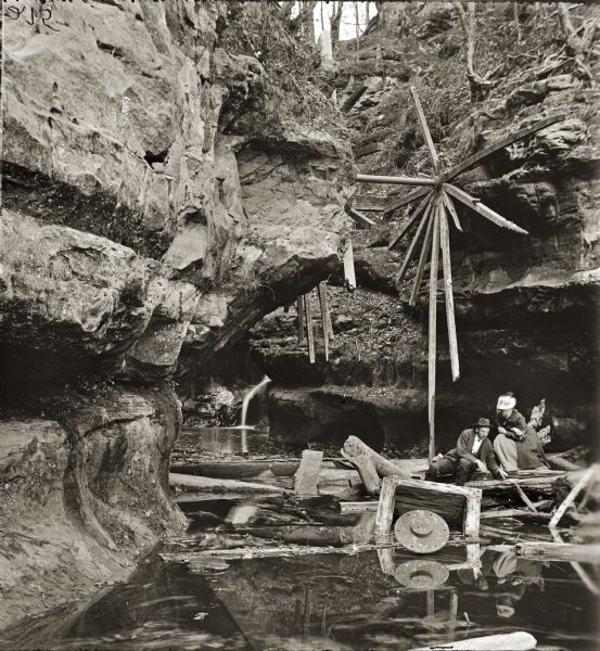 Man and woman sitting on logs near the water line at Pewit's Nest. Wooden debris on ground and attached to rock face. A small waterfall is in background.