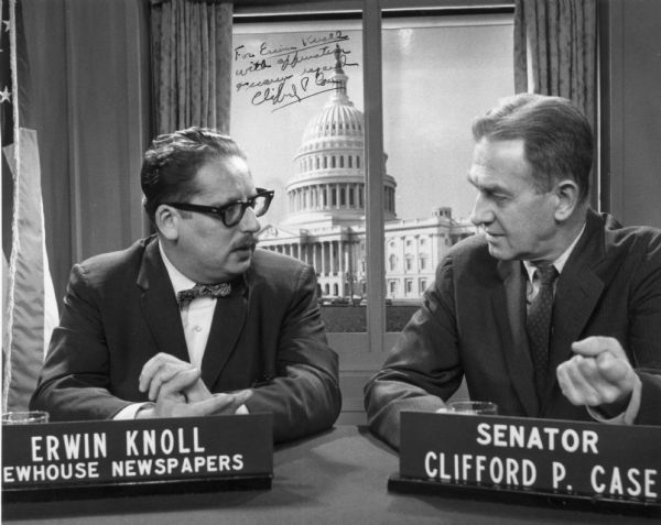 Erwin Knoll, then a reporter with the Newhouse Newspapers, interviewing Senator Clifford Case of New Jersey. Knoll later became the editor of the <i>Progressive Magazine</i>. They are sitting at a desk with nameplates in front of them, and behind them is a flag in front of curtains framing an image of the United States Capitol.
