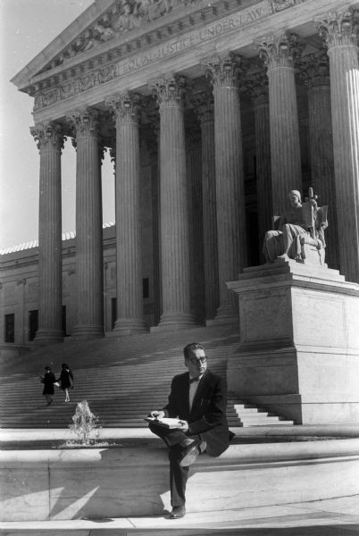 Journalist Erwin Knoll, wearing a suit and bow tie, posed sitting on a fountain in front of the entrance to the United States Supreme Court building during his years as a reporter. Knoll later became the editor of the <i>Progressive Magazine</i> in Madison, Wisconsin.