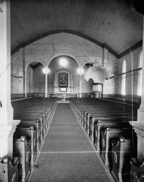 View of a church interior looking up the aisle between the pews and towards the altar. An acoustical hood or sounding board is over the pulpit, and the altarpiece includes a painting. The church's stone walls are exposed.