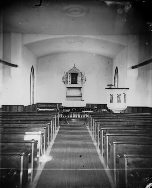 View of the interior of the Norwegian-American Lodi Church, looking up the aisle and towards the Acanthus design altar, which is surrounded by a wooden railing, and an organ. A painting hangs above the altar.