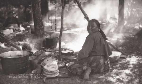 Winnebago (Ho-Chunk) Indian camp with woman seated on the ground making fry bread. There is a kettle over the fire, with  a tripod holding a pot for cooking.
