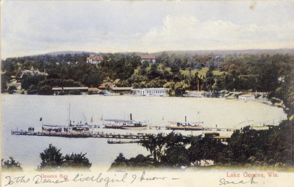 Elevated view of Lake Geneva Bay, with assorted homes and hotels, and boats docked at long piers. Caption reads: "Geneva Bay, Lake Geneva, Wis."