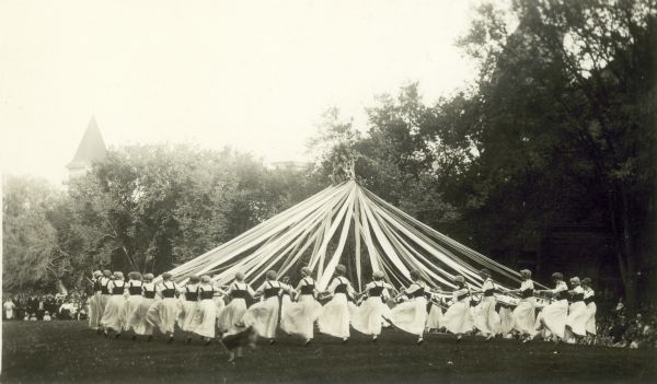 A group of women in white skirts and wearing caps are dancing around a tall maypole. A crowd is watching in the background, and a dog is running in front of the group on the grass.