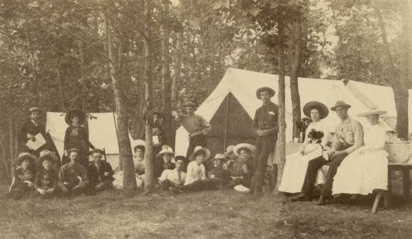 University of Wisconsin-Madison students and chaperons during a camping trip at Livesy Springs on the north shore of Lake Mendota. On the left side stands Professor King, of the Engineering Department, with his wife. Many of the women are holding or wearing sprays of flowers, and others are holding playing cards.