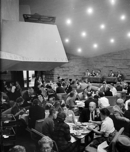 Interior view of the Auditorium of the Frank Lloyd Wright-designed First Unitarian Society Meeting House. The Wright furniture has been arranged to illustrate the multiplicity of uses of the space. A large group of people are eating at tables throughout the room.