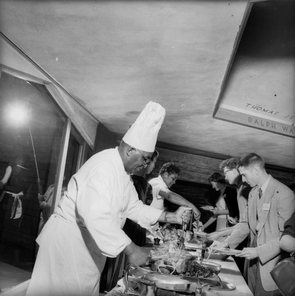 Carson Gulley, the well-known University of Wisconsin-Madison chef, serving a catered meal at the Frank Lloyd Wright-designed First Unitarian Society Meeting House. The hexagonal space designed to alleviate the impact of the low ceiling in this area of the building can be seen above the serving line. Two rows of names of Unitarian leaders decorate the hexagon. The top row of names was later painted over.