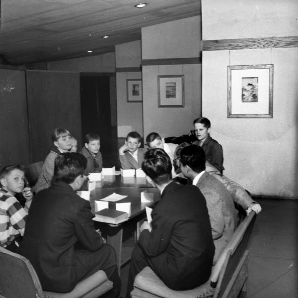 Children seated around a triangular table in the loggia (hallway) of the Frank Lloyd Wright-designed meeting house of the First Unitarian Society.  They appear to be playing a game. Some of the Japanese prints given to the Madison Unitarians by Wright can be seen hanging on the wall behind them.