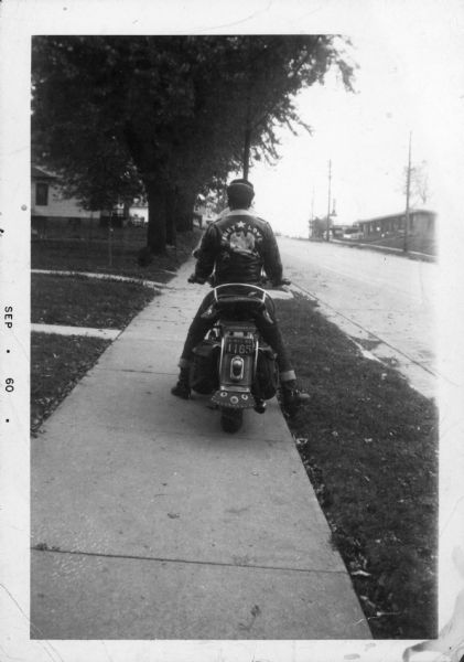 View from behind of Lewis Arms riding his 1946 Harley-Davidson  Flathead motorcycle on the sidewalk of the 1800 block of Beld Street. On the back of his jacket is the nickname "Honey Love."
