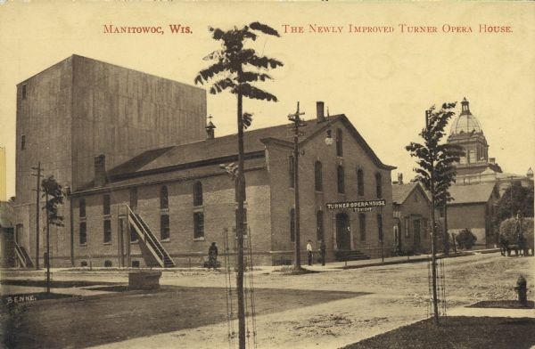 Exterior view of the Turner Opera Hose with the Manitowoc County Courthouse in the background. Horses pull a wagonload of hay at right. Caption reads: "The Newly Improved Turner Opera House, Manitowoc, Wis."