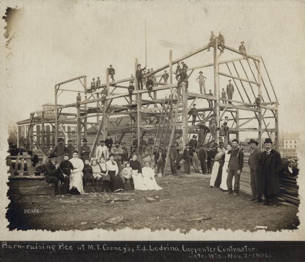 Group of people assembled in front of and on the frame of a barn being raised at M.T. Cooney's.