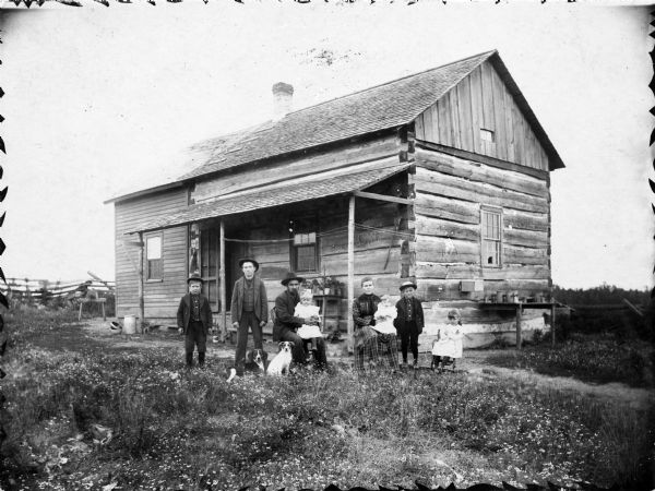 Husband and wife with their six children and two dogs posed in front of their log cabin home. There is a framed addition on one side of the structure and a split-rail fence in the background.