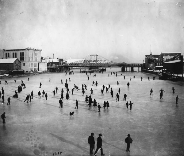 Elevated view of a winter scene with a large group of people skating on the frozen Manitowoc River between the 8th and 10th Street bridges. A dog trots across the ice near the bottom center and a tug boat is frozen along a pier on the right.