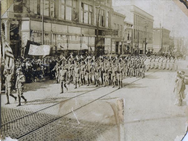 Soldiers parade with guns bearing American flags past Schuette Brothers clothing store at the corner of S. 8th and Jay Streets as spectators look on. There is a street car track running down the center of the brick-paved street.