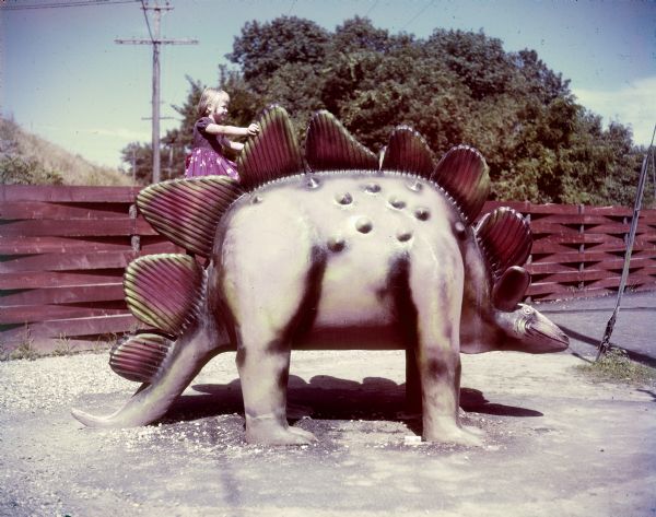 A girl climbs on the back of a model stegosaurus in Prehistoric Land, part of the Enchanted Forest in Wisconsin Dells. There is a wood fence of woven planks in the background.