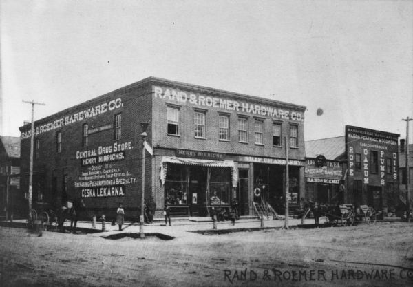 Exterior view from street of Rand & Roemer Hardware Co. and Henry Hinrich's Drug Store. Also visible are Rand & Roemer Ship Chandlery (nautical supply), Rand & Roemer Carriage and Hardware, and Rand & Roemer Hardware. There are several pedestrians on the sidewalk and horse-drawn vehicles on the street.