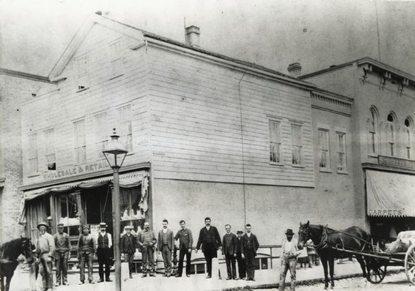 Men who worked for Schuette Brothers Store assembled in front of the store. There are horses on both sides of the photograph, and the one on the right is pulling a wagon.
