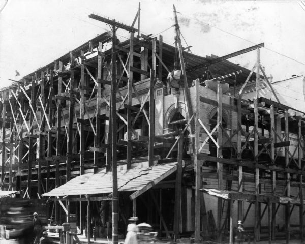 Construction crew raising a brick building. The structure is entirely surrounded by scaffolding. A large stone piece is being hoisted into place while a woman walks across the street in the foreground pushing a baby in a carriage.