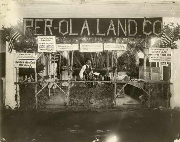 Man standing in a booth promoting the Per-Ola Land Company. There are several Forest County grown grains and crops on display in the booth to show the bounty of the available land. Signs in the booth advertise Kennan Silt Loam, prices paid for Forest County wood products, independence of farm life, home sites at Stone Lake, land for $30/acre. There are American flags on either side of the exhibit.