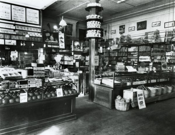 Interior view of Schuette Brothers grocery department. Among the items for sale are grapefruit, apples, bananas, dates, and figs. There is a Jell-O advertisment at the back of the store, and what appears to be a pulley system at the right.