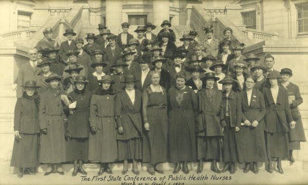 Group portrait taken on the stairs of the Wisconsin State Capitol of men and women attending the State Conference of Public Health Nurses. Most of the women wear long coats and hats.