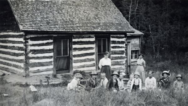 Theta Mead posed outside a log cabin schoolhouse with a teacher and her class. One of the girls has her arm around a dog.