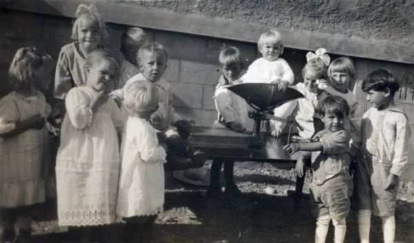 Group of small children gathered around a table outdoors near a building on which a toddler is being weighed using a baby scale.