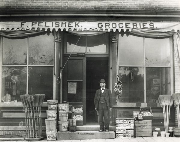 Frank Pelishek standing in front of his grocery store. Items on display in front of the store include brooms, candy, bananas, seed packets, and flower pots.