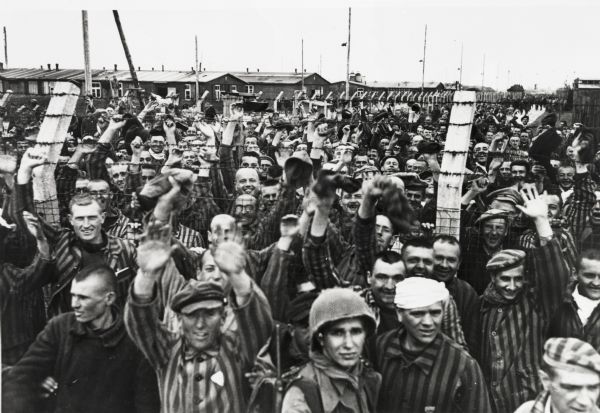 Prisoners, most wearing striped uniforms, on both sides of a barbed wire fence cheering at their liberation from the concentration camp by the United States Army. An American soldier stands in the foreground.